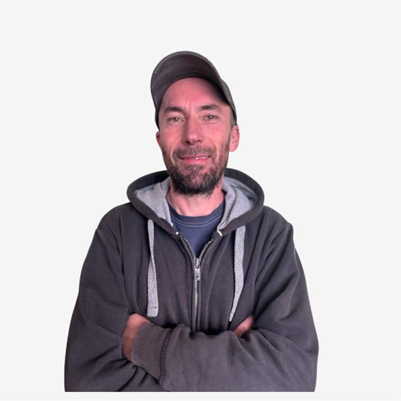 Portrait homme décontracté avec casquette et sweat à capuche Homme barbu d'âge moyen souriant, portant une casquette grise et un sweat à capuche zippé foncé, les bras croisés sur fond blanc.