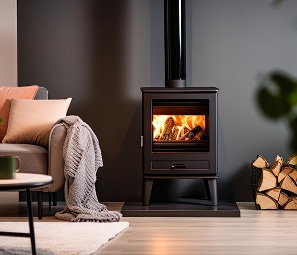 Poêle à Bois Design : Chaleur Moderne et Ambiance Cocooning Poêle à bois noir moderne avec feu vif, posé sur une plaque, à côté d'un canapé gris et d'un tas de bûches.